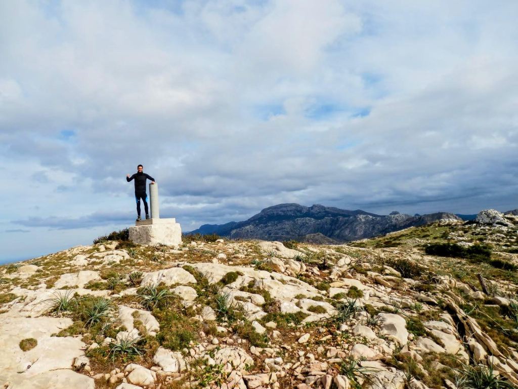 Persona en la cima del Puig de Ses Basses, posando al lado de un hito geodésico con un paisaje montañoso al fondo.