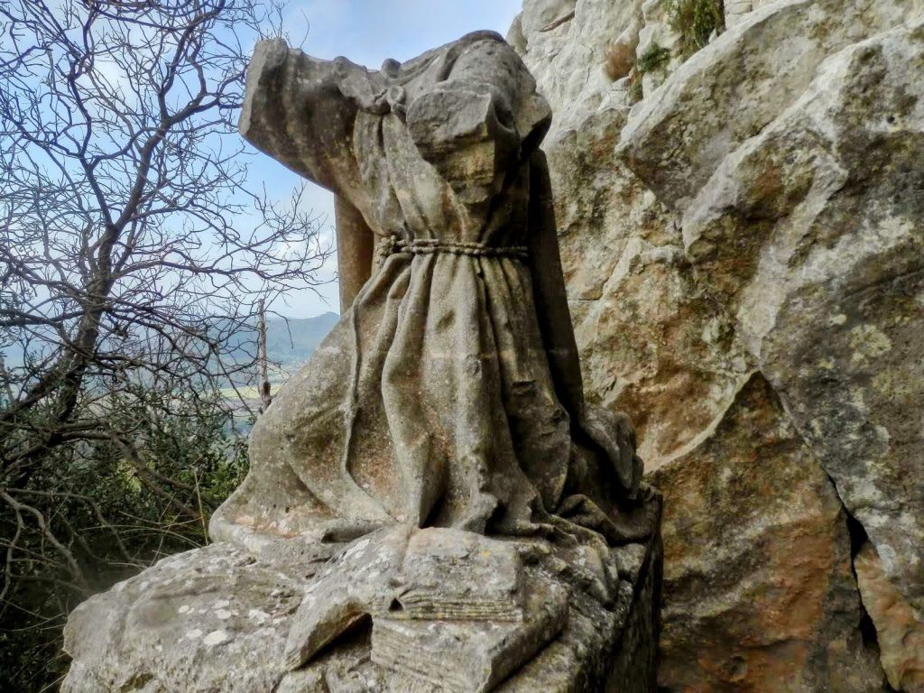 Estatua fragmentada del Beato Ramón Llull en un entorno rocoso, con vegetación en el fondo.