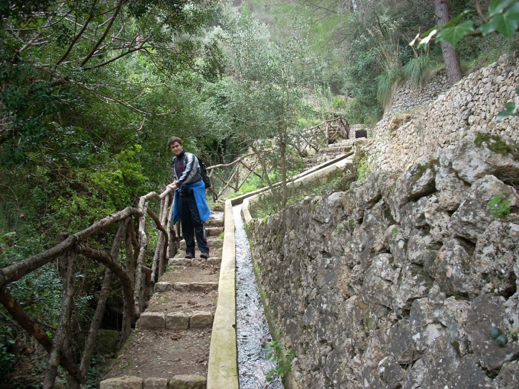 Sendero empedrado con barandilla de madera rodeado de vegetación, con una persona en la escena., en la Font de la Vila 