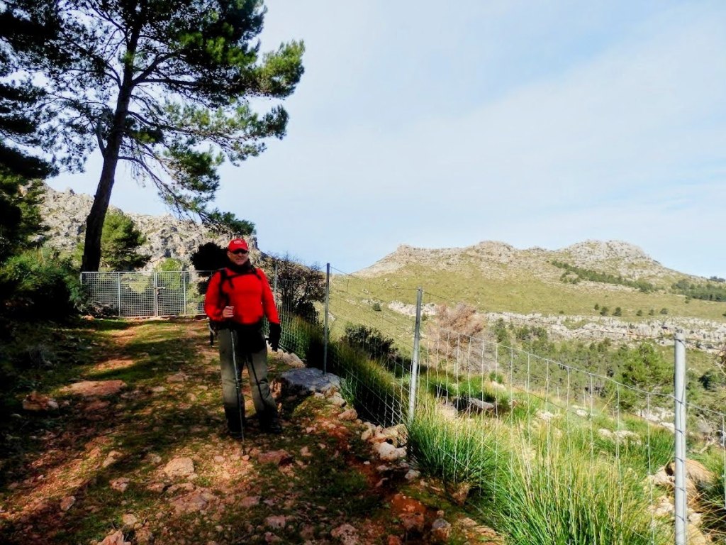 Un senderista vestido de rojo se detiene en un camino de tierra, rodeado de vegetación y montañas de fondo. Hay una valla metálica a un lado, y el cielo está parcialmente nublado.