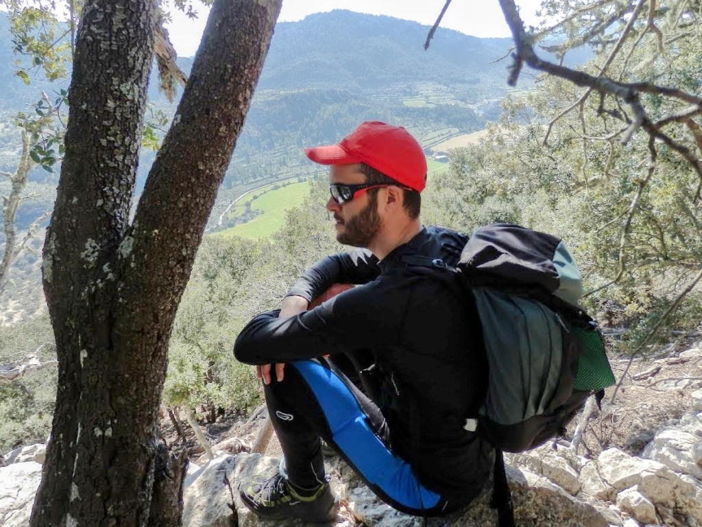 Hombre sentado sobre una roca, vestido con ropa de senderismo y mochila, mirando hacia el Valle de Orient, con árboles y campos verdes.