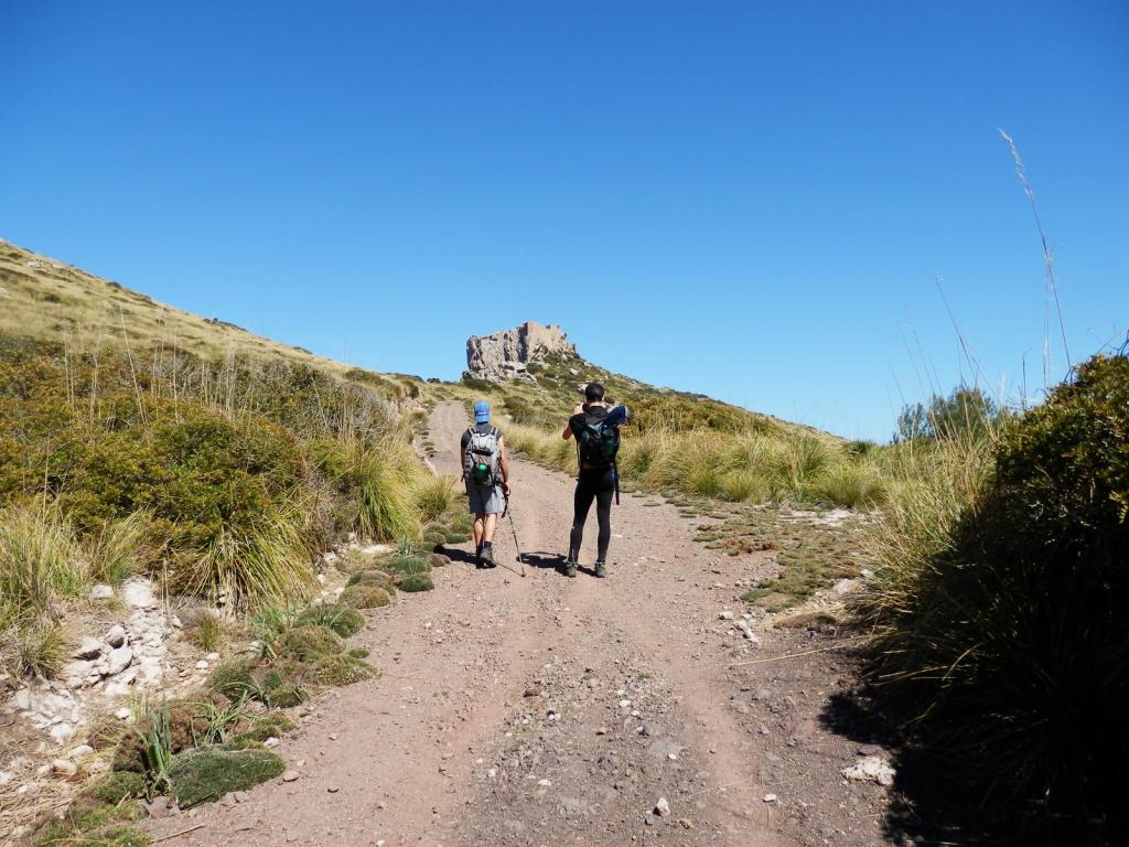 Dos senderistas caminando por un camino de tierra rodeado de naturaleza, con un paisaje montañoso al fondo, incluyendo ruinas de un castillo en la cima.