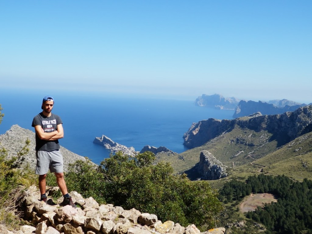 Hombre de pie en un paisaje montañoso, con vistas al mar y acantilados en el fondo, rodeado de vegetación.
