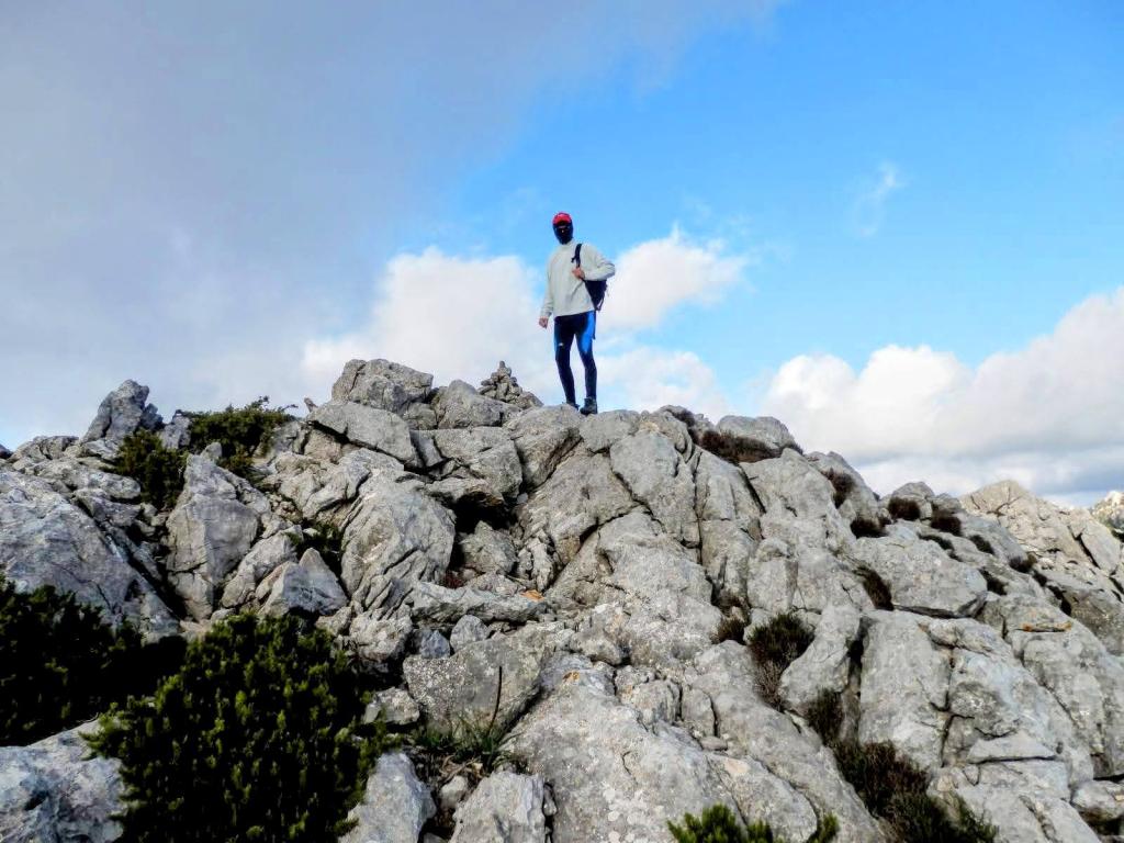 Persona de pie sobre la cima del Puig des Coll des Jou con un paisaje montañoso y un cielo nublado al fondo.