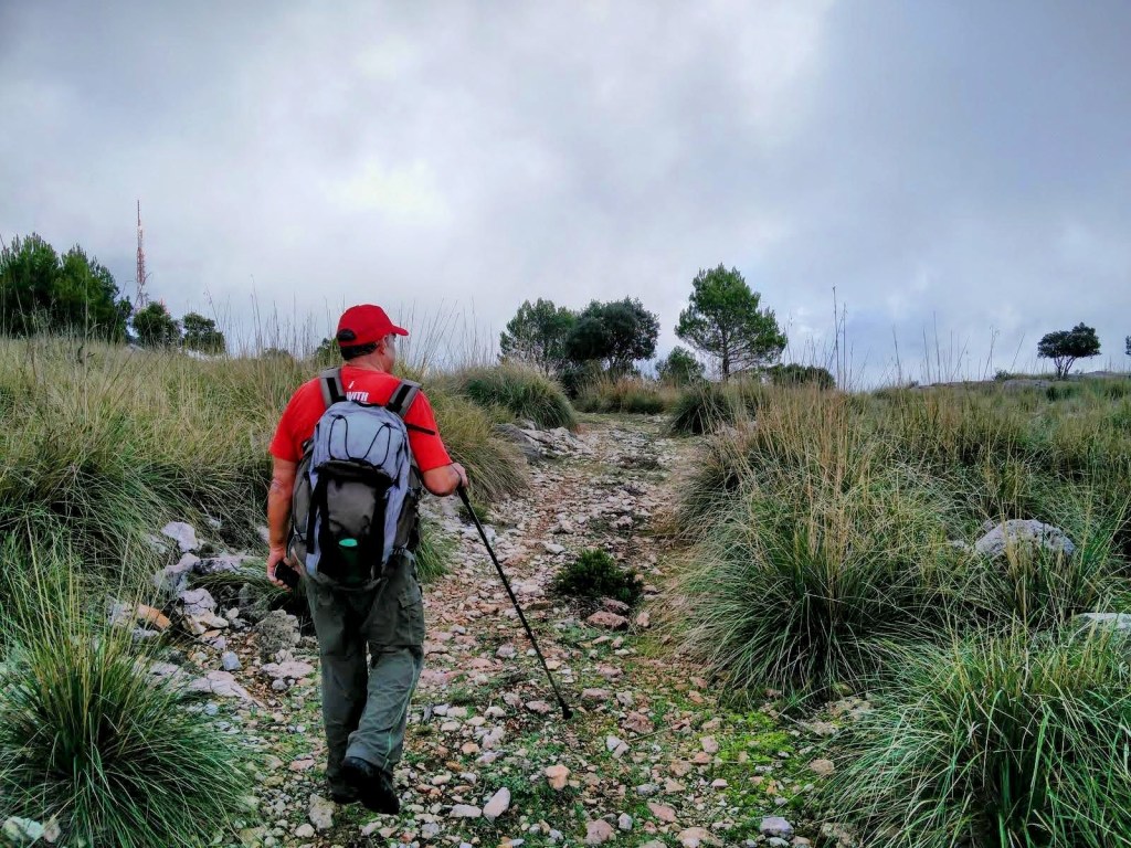 Una persona caminando por un sendero pedregoso en un paisaje de montaña, con vegetación alta y antenas visibles al fondo bajo un cielo nublado.