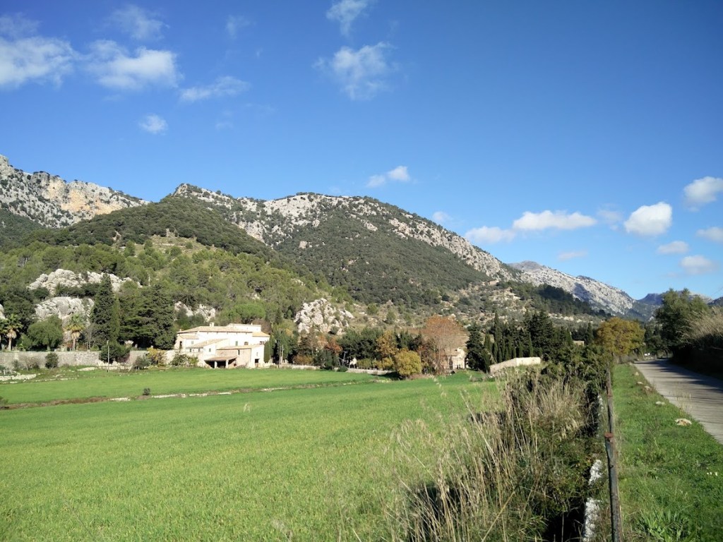 Vista de un paisaje montañoso en la ruta Puig des Coll des Jou, con campos verdes en primer plano y Son Vidal entre árboles.
