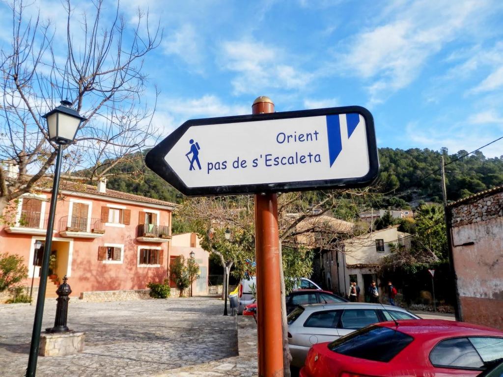 Señalización del camino hacia 'Orient' y 'Pas de s'Escaleta' en Alaró, Mallorca, con edificios y árboles de fondo.