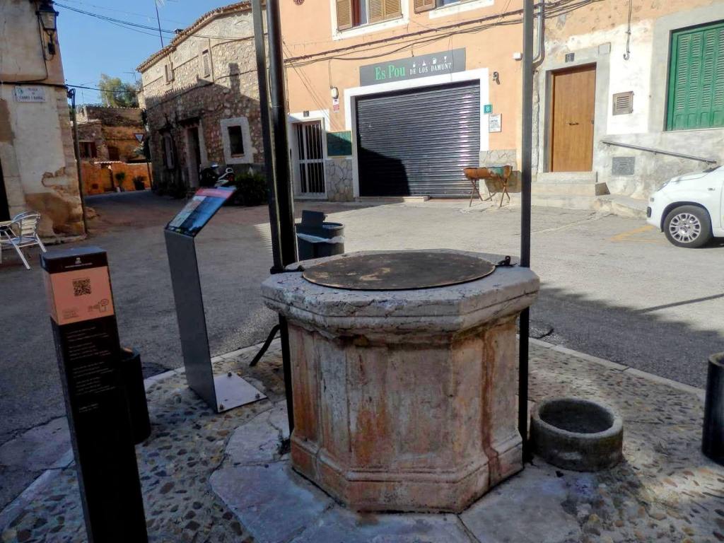 Vista de un pozo de piedra en  la Plaza de Cabrit i Bassa, con señales informativas a su alrededor y edificios de fondo.