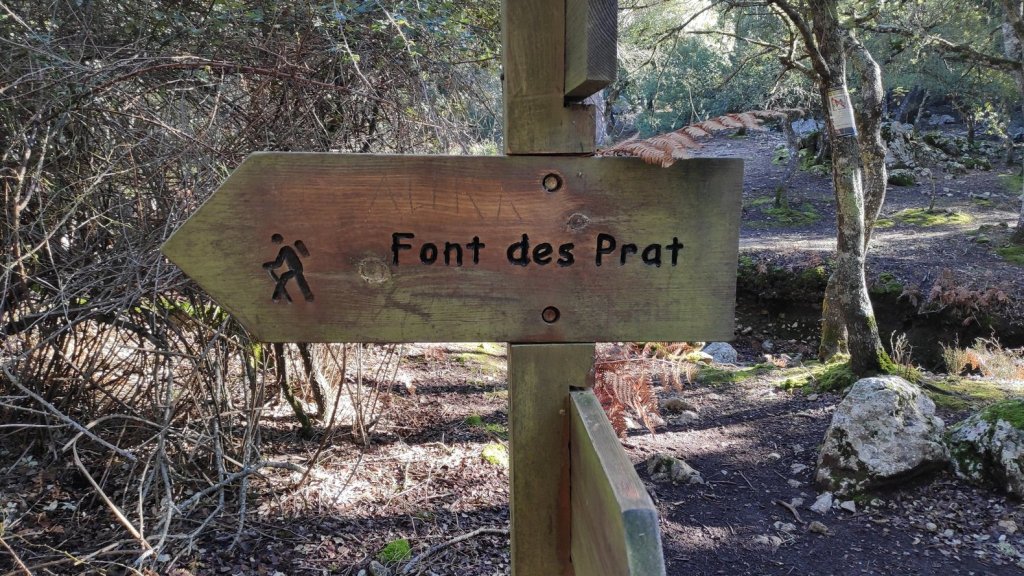 Señal de madera indicando la dirección hacia 'Font des Prat', rodeada de vegetación y naturaleza.