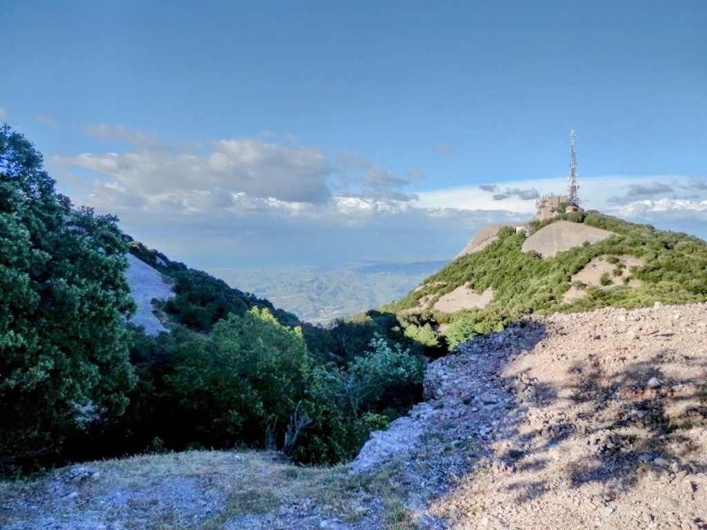 Vista panorámica del Serrat del Moro con su cumbre y una torre de telecomunicaciones en la cima, rodeada de vegetación y un cielo despejado.