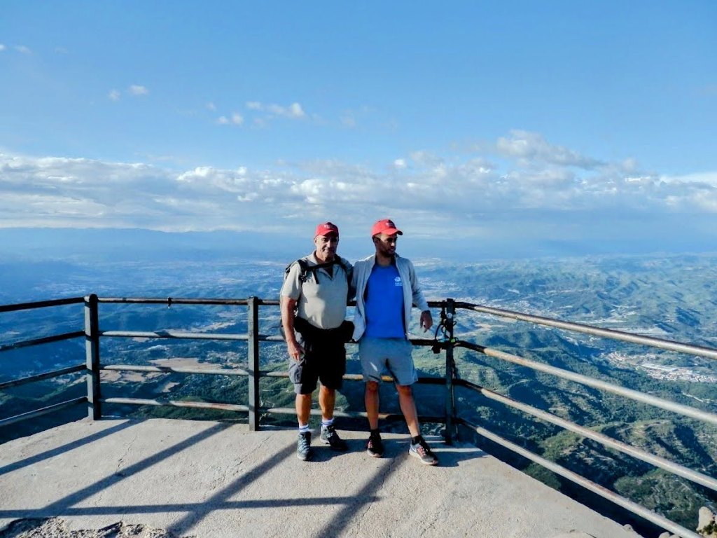 Dos personas de pie en un mirador en la cima de Sant Jeroni, con vistas panorámicas de las montañas y el paisaje de Montserrat.