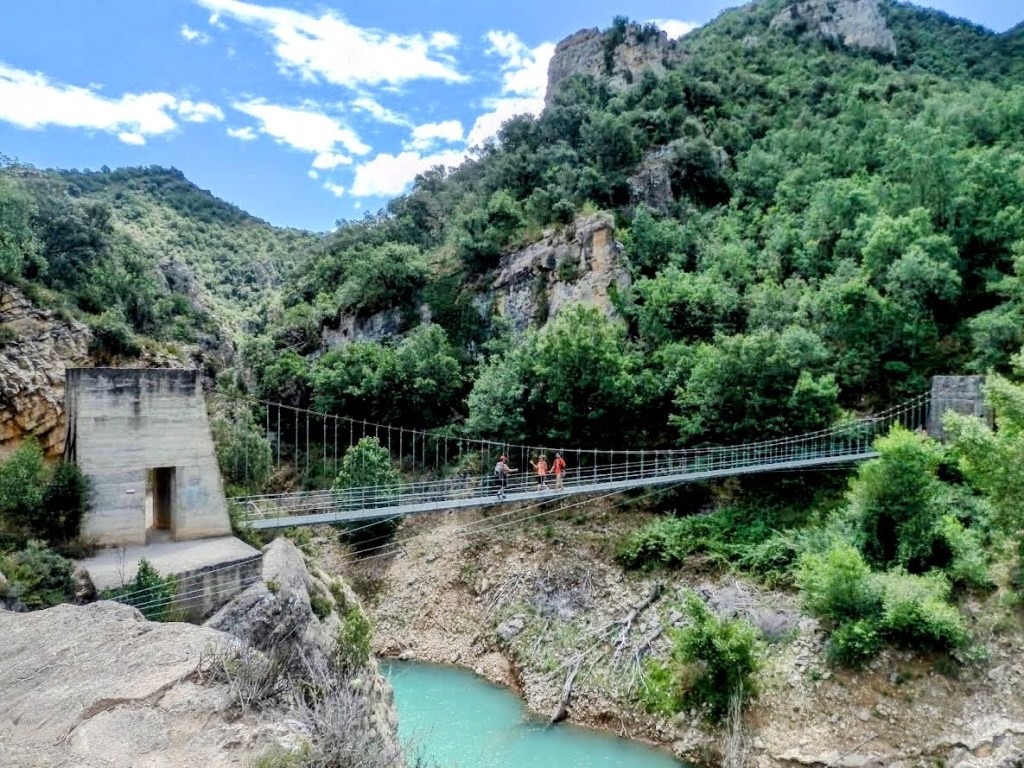 Puente colgante en el desfiladero de Mont-Rebei, rodeado de vegetación y montañas, con personas cruzando sobre un río de agua clara.