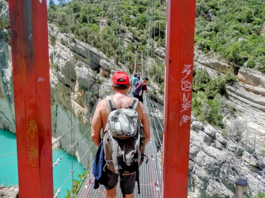 Persona cruzando el Puente colgante del Congost del Seguer, rodeado de paredes rocosas y vegetación, con agua verde turquesa visible abajo.