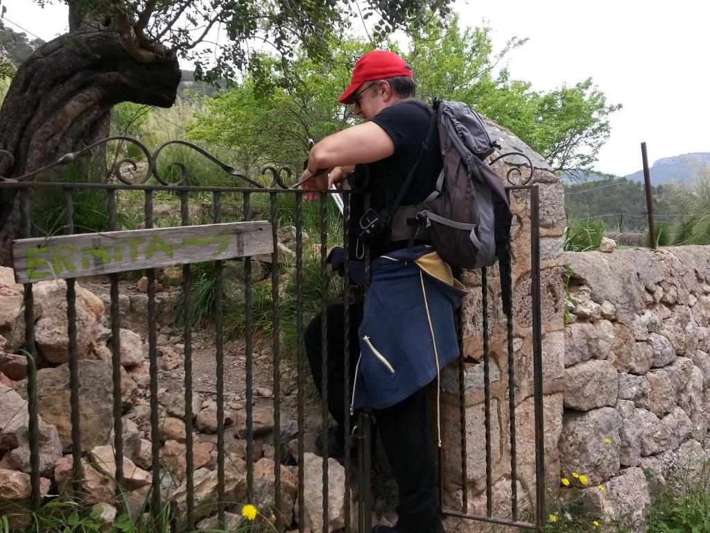 Una persona encaramada en una puerta de hierro con la inscripción 'Ermita' mientras explora un sendero con vegetación alrededor.