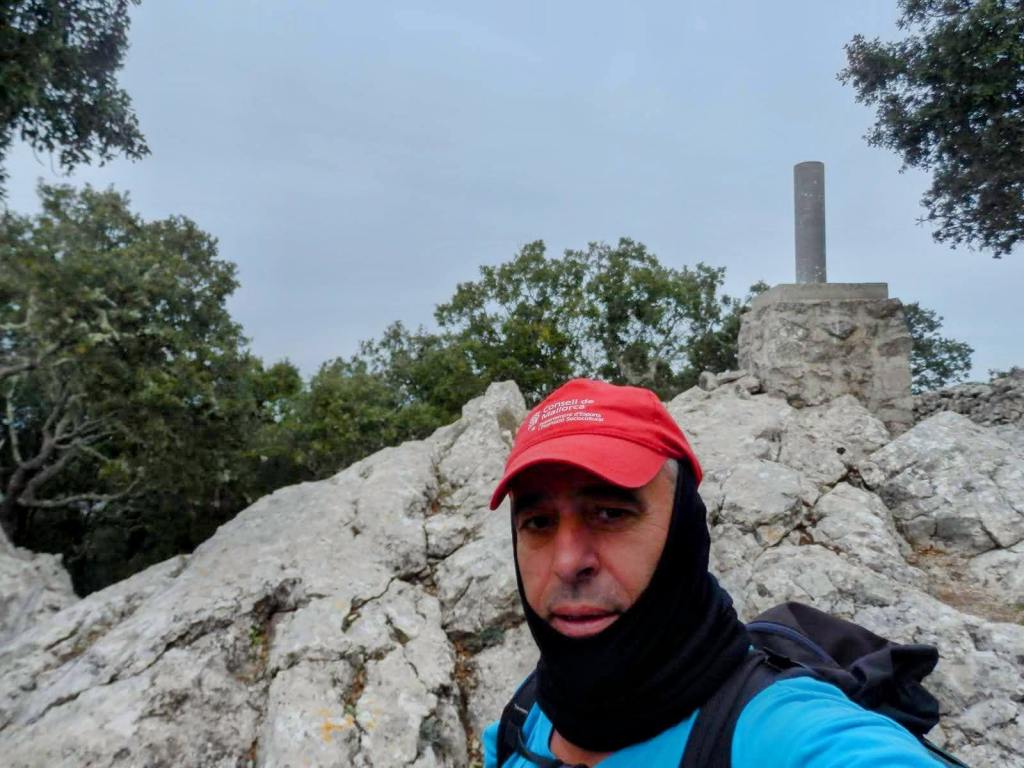 Autorretrato de un senderista en la cima de la Fita del Ram, con un vértice geodésico visible al fondo, rodeado de rocas y vegetación.
