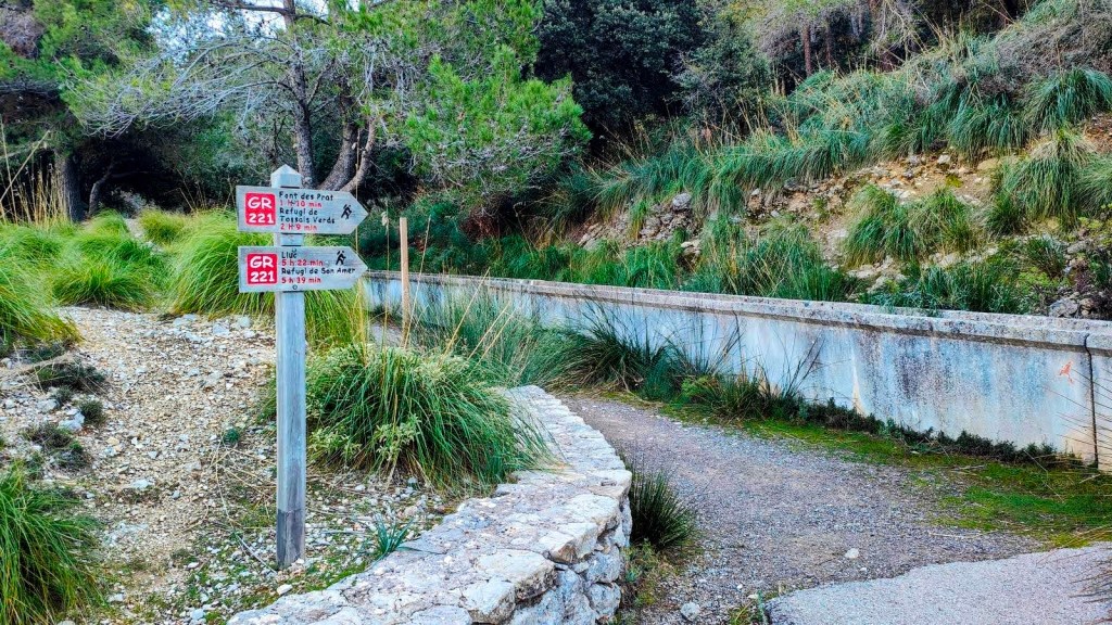 Señalización del sendero GR 221 en un área natural, con un camino de tierra, vegetación alrededor y un canal de agua visible.