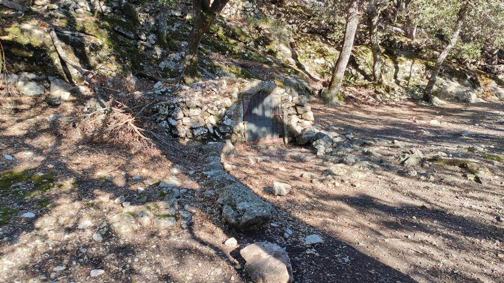 Entrada de la Font des Prat, parcialmente oculta entre árboles, con un marco metálico que indica una puerta cerrada, en un entorno natural con suelo de tierra y rocas.