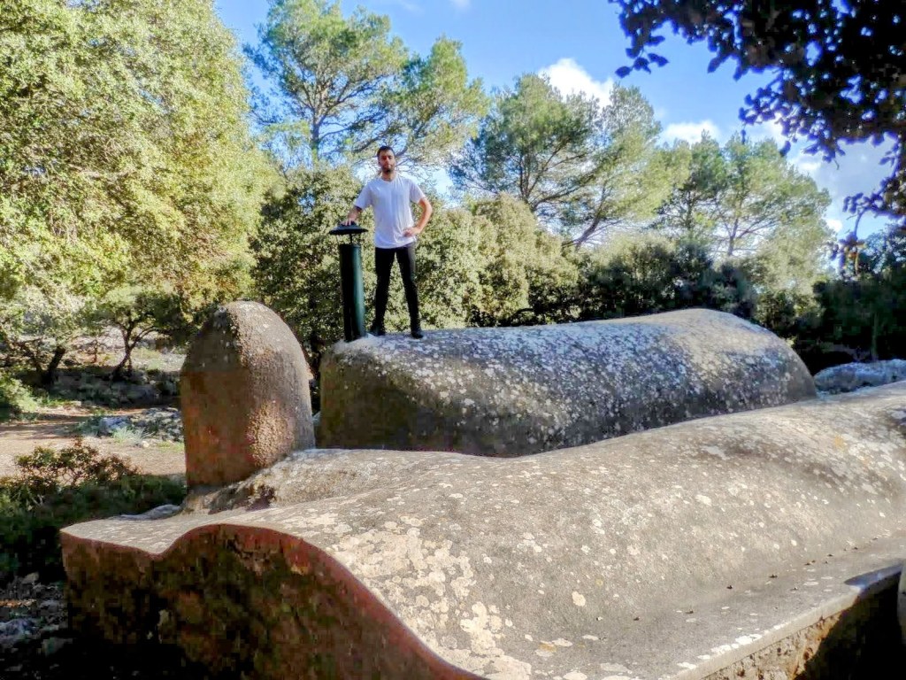 Persona de pie sobre el Aljibe de Mestre Pericó Barbut, en un entorno boscoso, con árboles de hoja perenne al fondo.