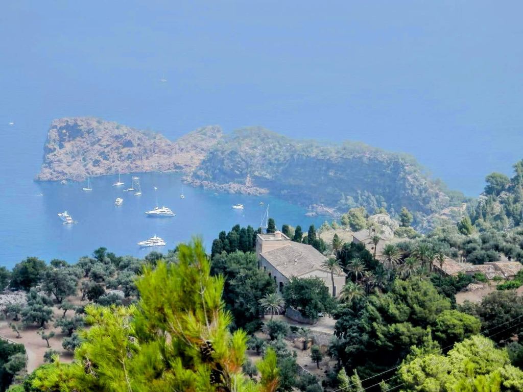 Vista panorámica de un paisaje costero con un acantilado, un edificio con un tejado de dos aguas, árboles y varias embarcaciones en el agua.