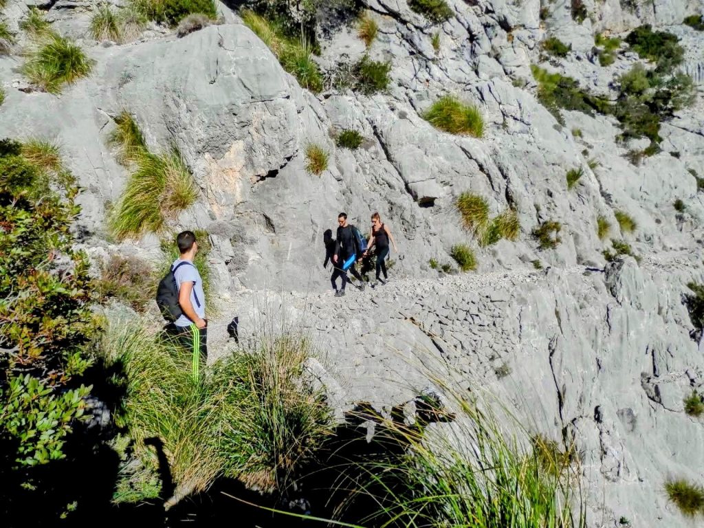Grupo de tres personas caminando por el Pas de ses Cases rodeado de rocas y vegetación en la Ruta La Cima del Puig Roig en Mallorca.
