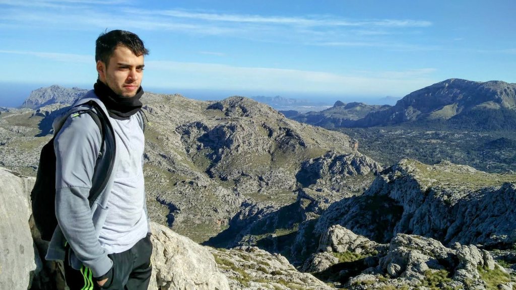 Hombre de pie en la cima del Puig Roig, con vistas panorámicas de montañas y valles a su alrededor, cielo despejado en el fondo.