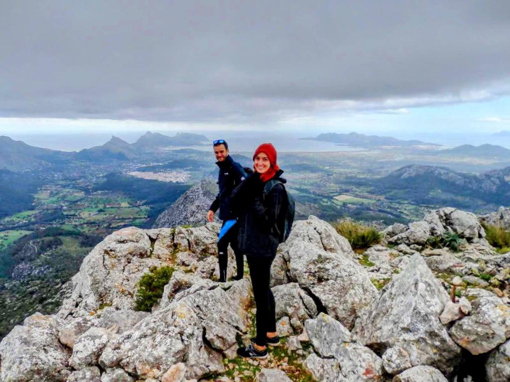 Dos personas en la cima de la Cuculla de Fartàritx, rodeadas de rocas y un paisaje montañoso de fondo, con un cielo nublado.