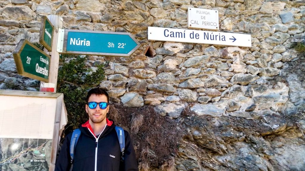 Un senderista con gafas de sol se encuentra frente a un panel de señalización en la ruta hacia Núria, con un fondo de pared de piedra.