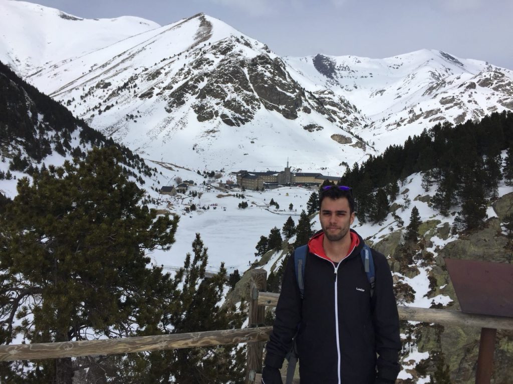 Caminante en el Mirador Cruz d’en Riba, frente a un paisaje montañoso cubierto de nieve, con el Santuario de Núria visible en el fondo.