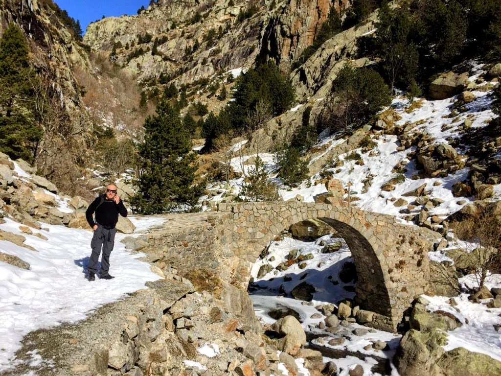 Un hombre de pie junto al Puente de Cremal en un paisaje montañoso cubierto de nieve, rodeado de árboles y rocas.