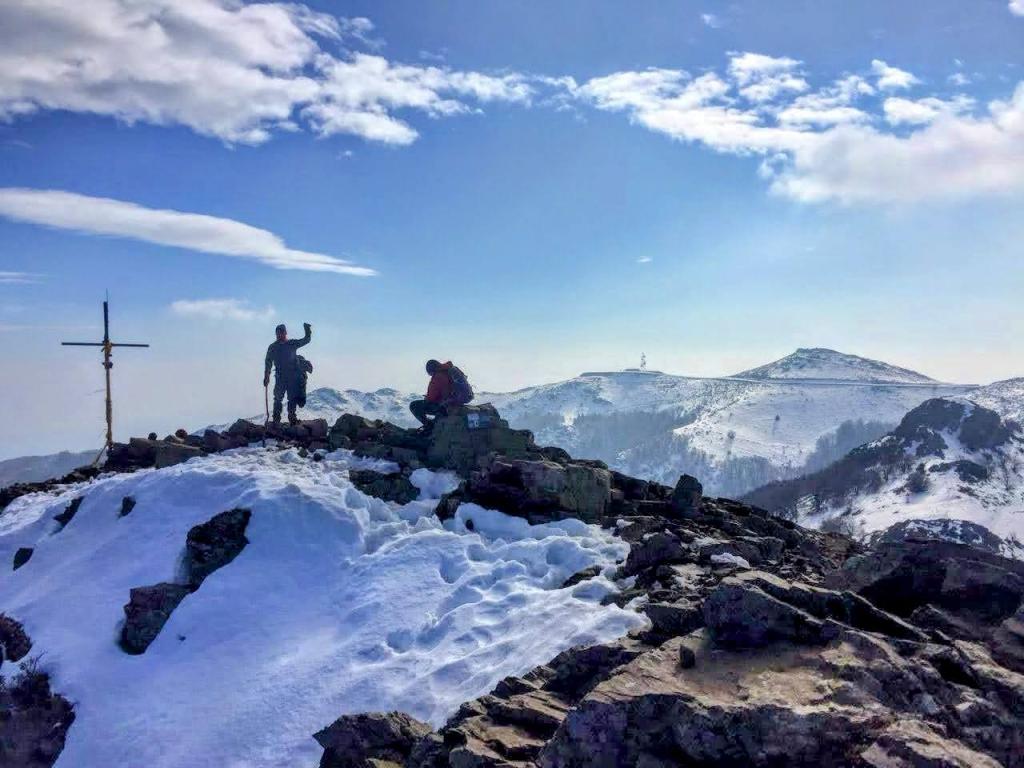 Dos personas en la cima nevada de Les Agudes, una de ellas levantando el brazo en señal de victoria, frente a un paisaje montañoso bajo un cielo azul con nubes.