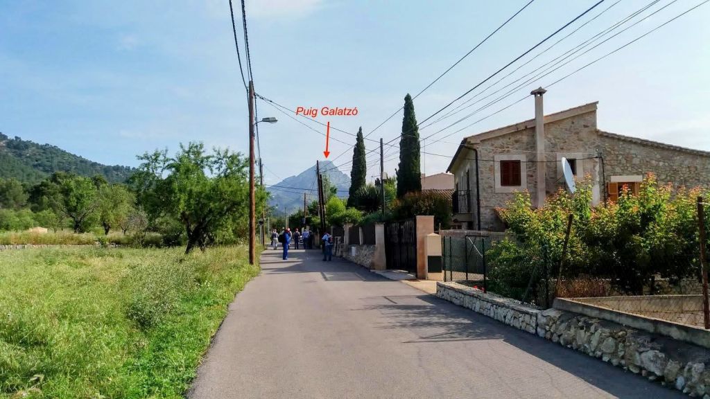 Imagén de la ruta hacia las Casas de Galatzó, mostrando una calle con casas y vegetación al lado. Al fondo, se puede ver la montaña Puig Galatzó. Varias personas caminan por la acera.