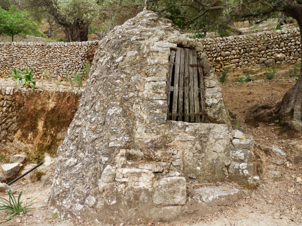 Estructura de piedra con puerta de madera, rodeada de vegetación y muros de piedra en el fondo.