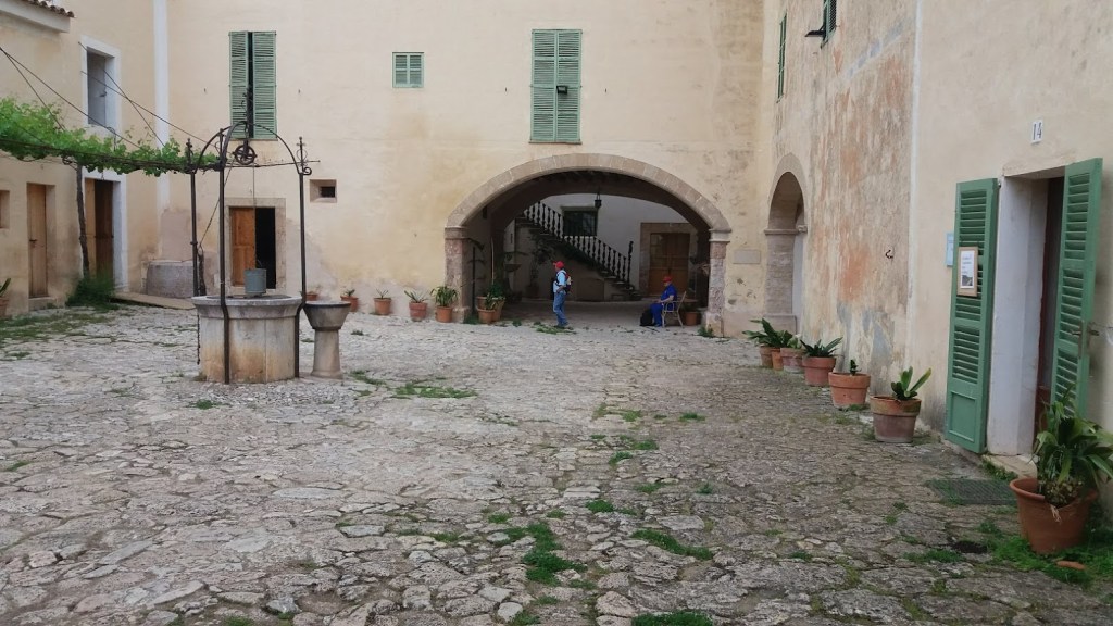 Plaza en el interior de las Casas de Galatzó, con un pozo en el centro, rodeada de edificios de piedra y plantas en macetas.