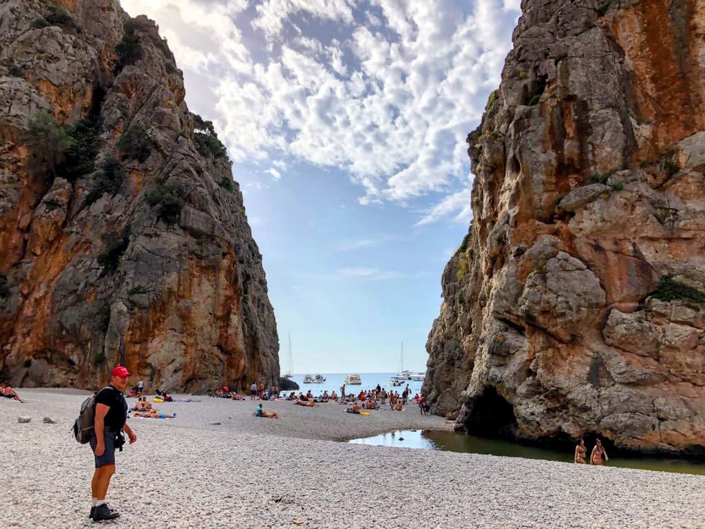 Vista de la desembocadura del Torrent de Pareis en Sa Calobra, con altos murallones de roca a ambos lados y un grupo de personas disfrutando en la playa de piedras blancas.
