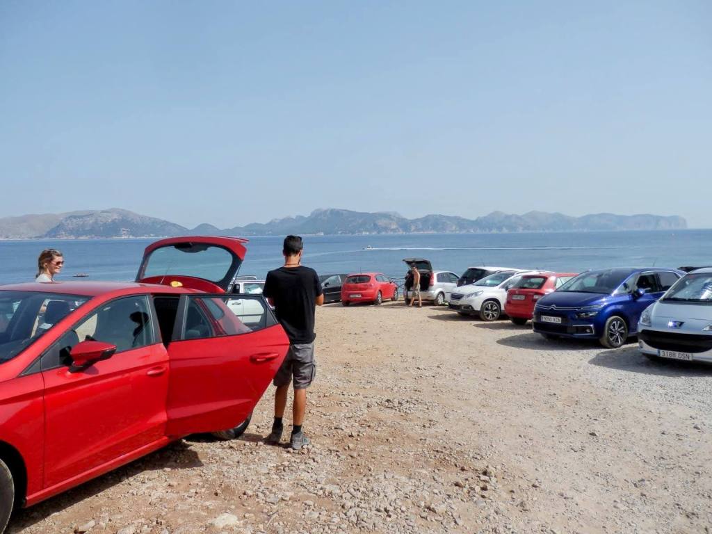 Vista de un aparcamiento junto al mar, con varios coches estacionados, incluyendo un coche rojo. Dos personas están de pie cerca de un vehículo, mientras el fondo muestra montañas frente al agua.