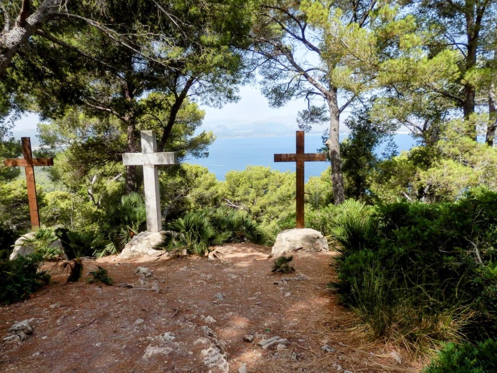 Tres cruces de madera y piedra en un entorno natural con vistas al mar, rodeadas de árboles y vegetación.