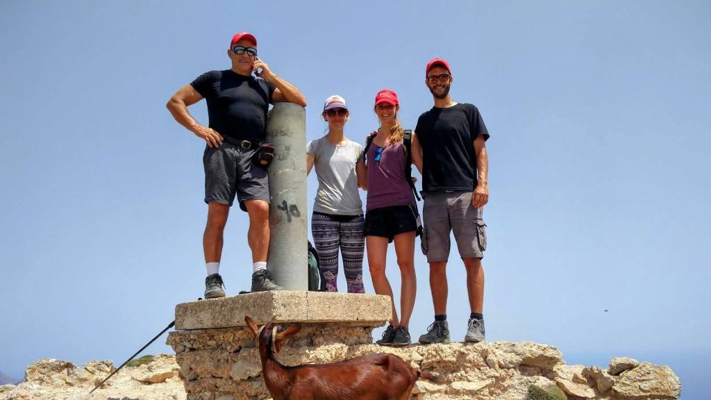 Grupo de cuatro personas en la cima de la Atalaya de la Victória, junto a un hito de piedra, con un cabra en primer plano y un cielo despejado de fondo.