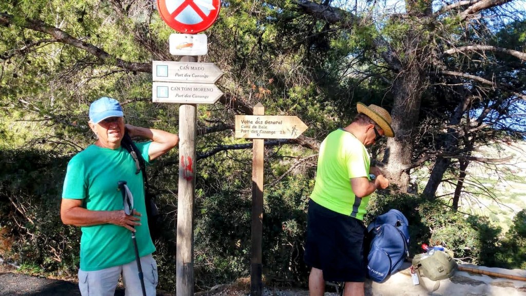 Dos hombres en un sendero al aire libre, cerca de señales informativas para excursionistas, rodeados de vegetación. Uno lleva una camiseta verde y sostiene un bastón de trekking, mientras que el otro usa una camiseta amarilla y está revisando su mochila.