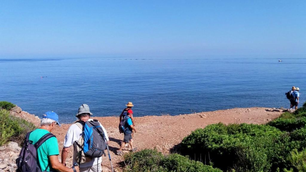 Grupo de caminantes con mochilas y gorros, caminando hacia la playa de Son Bunyola, con mar tranquilo y cielo despejado al fondo.