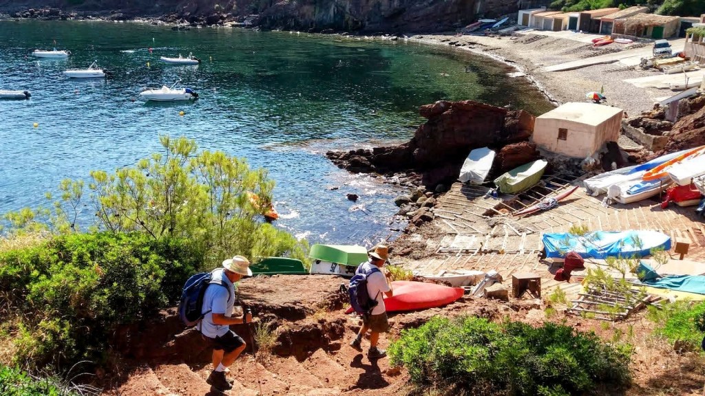 Dos senderistas descendiendo por un camino hacia una playa en el Puerto de Canonge, con botes y una desembocadura visible en el fondo.