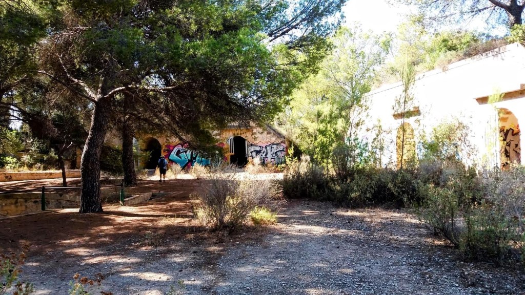 Ruinas de una antigua base militar, rodeadas de vegetación y árboles, con grafitis en las paredes. Dos personas caminan por un sendero cercano.