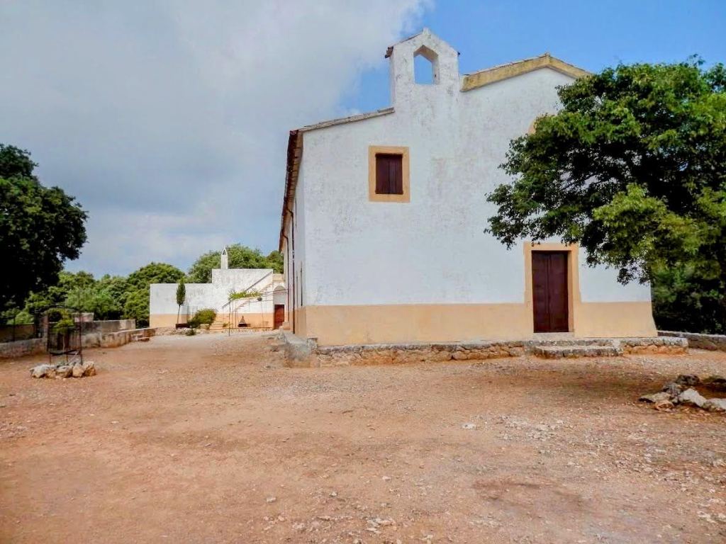 Vista exterior de la Ermita de Maristella, una construcción blanca con ventanas de madera, rodeada de terreno y árboles en un día nublado.