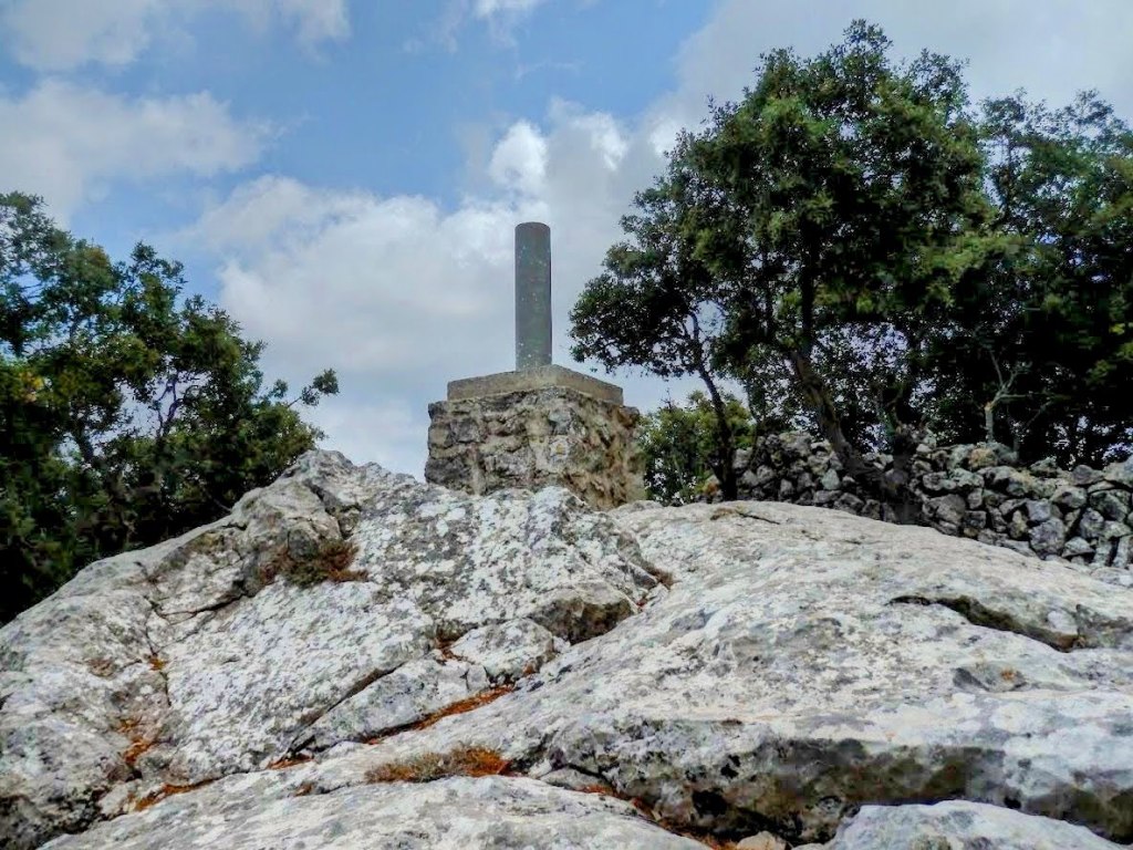 Vista de la cima de la Fita del Ram, con un vértice geodésico sobre una cordillera de rocas y árboles alrededor, bajo un cielo nublado.