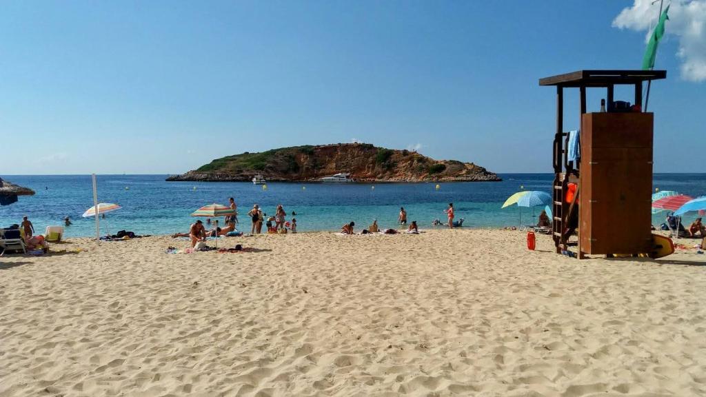 Playa con arena dorada y familias disfrutando del sol, sombrillas de colores y un salvavidas en la orilla, con una isla pequeña visible al fondo.