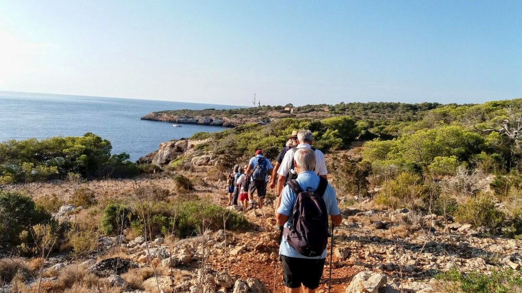 Grupo de senderistas caminando por un sendero costero cerca de Cala Figuera, rodeados de vegetación y vistas al mar.