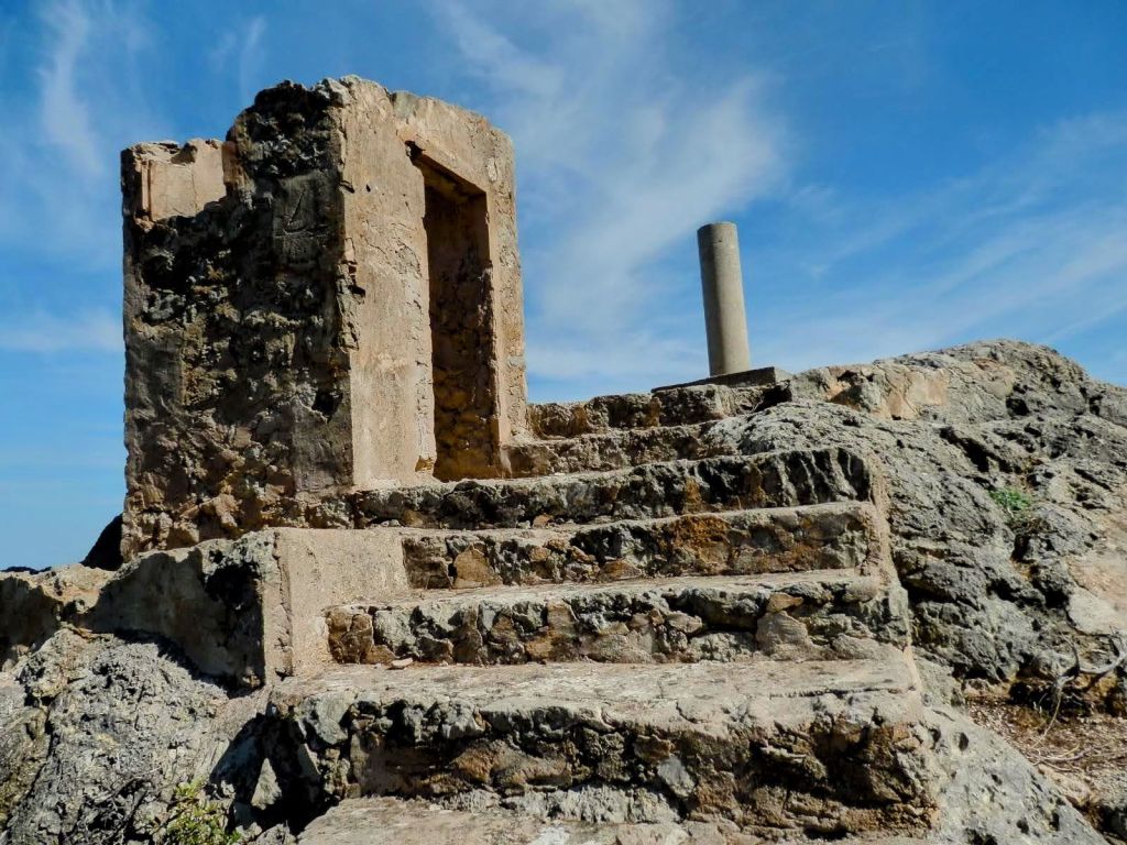 Estructura en ruinas con escaleras de piedra en la cima del Puig de ses Planes, iluminada por un cielo azul con nubes suaves.