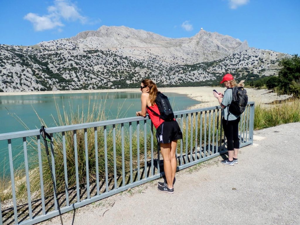 Dos personas observando el Embalse de Cúber rodeado de montañas, con una barandilla de metal entre ellas y el agua.