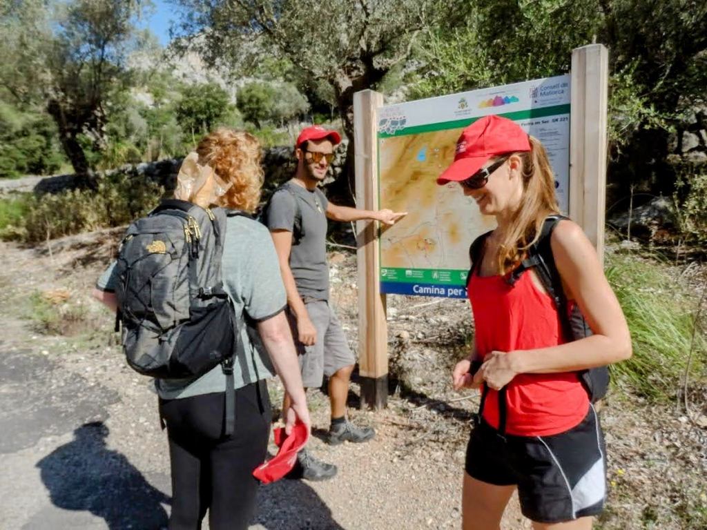 Tres personas en un sendero al aire libre, analizando un mapa de la ruta Tossals Verds. Dos mujeres, una con mochila y camisa deportiva roja, y un hombre con una camiseta gris, están de pie frente al cartel con información sobre el sendero, mientras la otra mujer observa con interés.