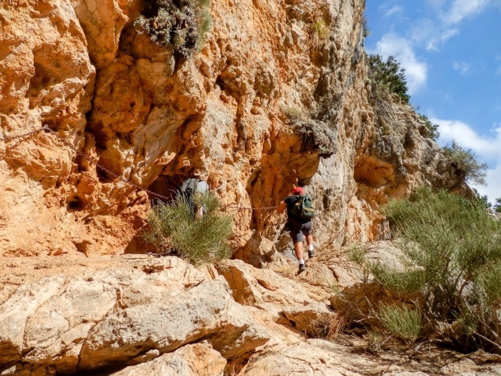 Escalador atravesando el Paso de Llis, con cadena de ayuda en la Ruta Tossals Verds por los Túneles, rodeado de vegetación.