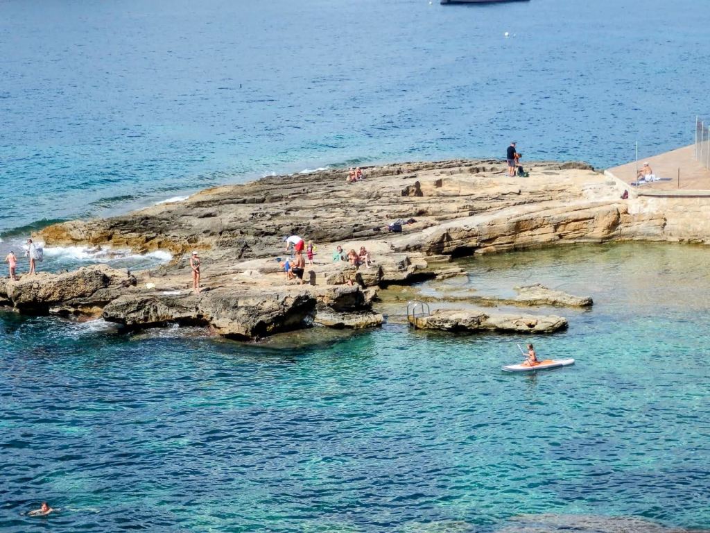 Vista de Cala es Conills con formaciones rocosas y personas disfrutando a lo largo de la orilla, algunas nadando y otras en una tabla de paddle surf, con agua cristalina y un mar tranquilo.